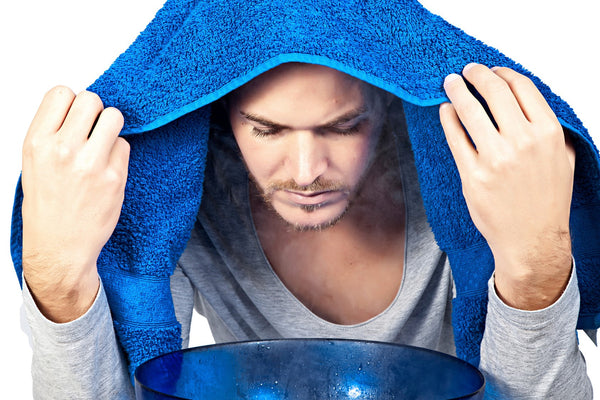 A man sits holding a blue towel over his head and blue bowl on the table in front of him with boiling water